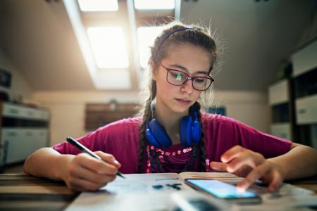 Teenage girl doing homework at home while using her smartphone. 